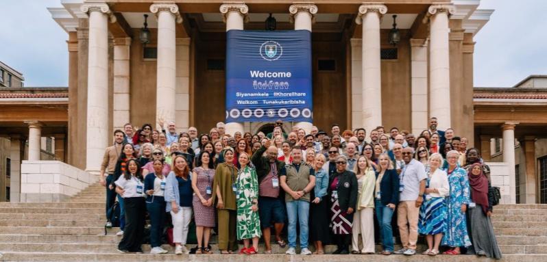 Image shows a group of people celebrating on steps, under a sign that reads the name of the conference they are attending.